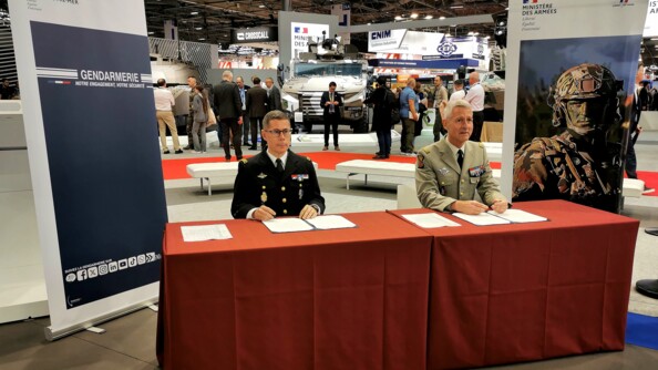 Dans une grande salle, assis à une table avec une nappe rouge, entourée de deux kakemono gendarmerie, le major général de la gendarmerie à gauche et le major général de l'armée de terre à droite signe un papier renouvellant ainsi l'accord de coopération.