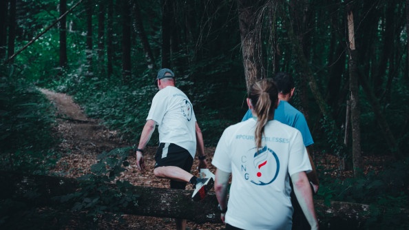 Trois personnes de dos sont dans un bois pour participer à une course d'orientation. Deux ont des t-shirt blanc et un, un t-shirt bleu clair.