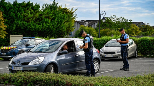 Sur un petit parking, deux gendarmes mobiles, sur la partie droite de l'image, procédent au contrôle d'une conductrice d'un véhicule léger gris.