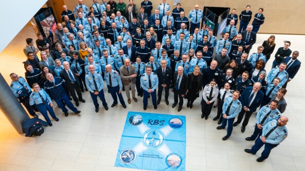 Dans un vaste espace intérieur, de nombreux gendarmes en tenue formant un demi-cercle sont photographiés par en haut. Tous lèvent la tête en direction de l'objectif. Au sol est disposé un grand tissu rectangulaire carré de couleur bleu ciel sur lequel il est inscrit en gros, en bleu marine RBS.