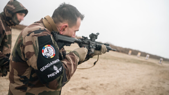 De trois quart arrière droit, un prévôt arborant au bras droit le brassard noir de la gendarmerie prévôtale s'entraîne au tir avec une arme d'épaule, sur un terrain sableux. Derrière lui, un militaire le guide
