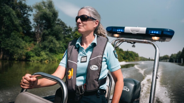 Une femme gendarme se tient debout, sur une embracation légère, avançant sur un fleuve. Equipée d'un gilet de sauvetage et de lunettes de soleil noires, elle se tient à l'armature métllique du siège situé devant elle.