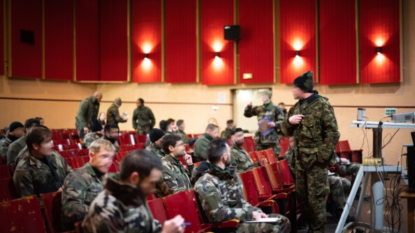 Les stagiaires sont dans un amphithéâtre rouge où ils écoutent un formateur.