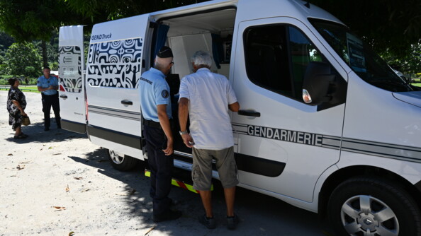 Devant le van blanc, aménagé, un gendarme échange avec un habitant en chemise blanche.