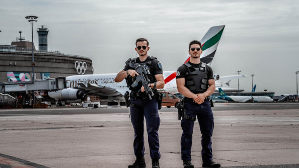 Deux gendarmes assurent la sécurité d'un aéroport. Ils se tiennent debout sur le tarmac. On aperçoit un avion ainsi qu'un bâiment gris circulaire portant sur la façade le sigle des JOP. L'un des deux gendarmes tient un fusil dans les mains.