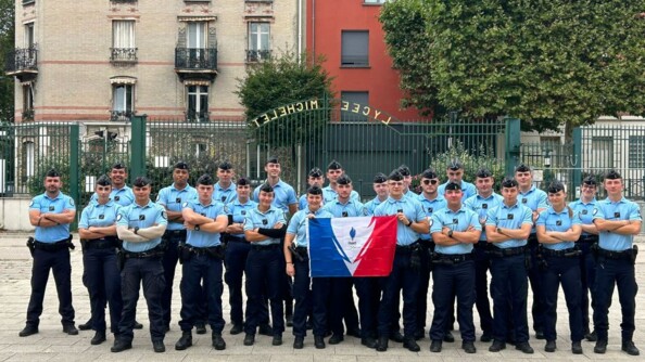 Un groupe de jeunes élèves-gendarmes pose avec le drapeau tricolore, sur une place, devant les grilles en fer forgé de couleur verte d'un lycée.