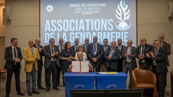 Au premier plan, le directeur général de la gendarmerie, assis derrière deux tables drapées d'une toile bleue portant la mention AMPHI apposée sur la flamme gendarmerie. Celui-ci tient un parapheur ouvert face à l'objectif, montrant plusieurs signatures sur deux pages côte à côte. Derrière lui, 17 personnes (15 hommes et deux femmes) en civil sont alignées en train d'applaudir. En arrière plan, sur grand écran, la mention au centre "ASSOCIATIONS DE LA GENDARMERIE" avec le logo de la flamme portée au creux de deux mains, avec un liseré bleu blanc rouge flottant en travers. En haut de l'image, la mention "Journée des Associations - lundi 24 juin 2024 - DGGN - Issy-les-Moulineaux - Amphi Moncey".