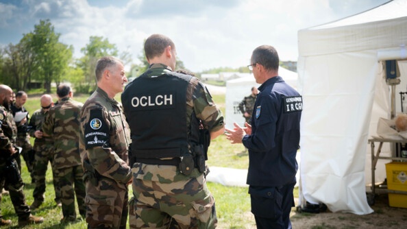 Trois gendarmes échangent, à gauche un prévôt de profil, en treillis, mains sur le ceinturon, avec le brassard de la gendarmerie prévôtale; au centre de dos, en treillis avec chasuble noire avec mention "OCLCH" dans le dos, un gendarme de l'OCLCH; à droite, de trois quart arrière en combinaison bleue, parlant avec les mains, un scientifique de l'IRCGN avec la mention "Gendarmerie IRCGN" dans le dos
