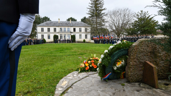 Au premier plan, une gerbe de fleurs et les jambes d'un gendarme au garde-à-vous. En arrière-plan, tout autour de la place d'armes en herbe, les troupes au garde-à-vous.