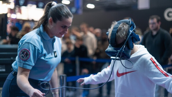 Une femme gendarme en tenue se tient aux côtés d'une jeune garçon d'une dizaine d'années. L'enfant porte un casque de réalité virtuelle devant les yeux, qui est relié par un fil à la main de la gendarme qui giude l'enfant