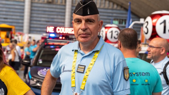 Un gendarme, vêtu d'un polo bleu ciel, et d'un tour de cou jaune portant la mention "Tour de France" au bout duquel est accroché un badge, se tient de bout, face à l'objectif. Derrière lui, on distingue une voiture et de nombreuses personnes regroupées dans un hangar.