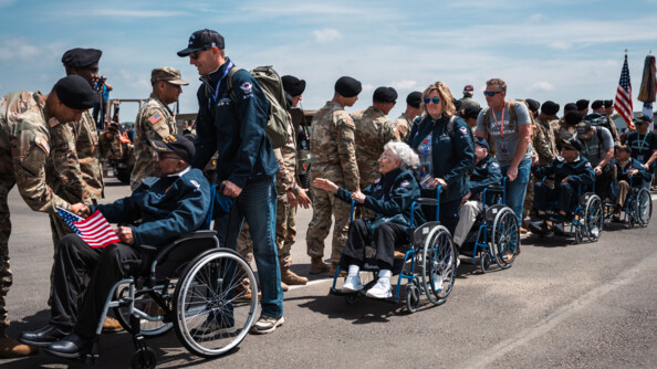 5 vétérans de la 2e guerre mondaile poussés en fauteuil roulant les uns derrière les autres serrent la main de militaires de l'armée américaine sur un tarmac