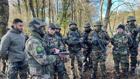 Neuf gendarmes en treillis dont un général quatre étoiles, major général de la gendarmerie nationale et plusieurs élèves grimés et porteurs d'un FAMAS écoutent l'un d'eux faisant un briefing
