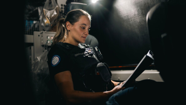 Une jeune femme gendarme, vêtue d'une tenue noire et d'un gilet par-balles, se tient dans un véhicule. Elle observe un document, qu'elle tient dans ses mains.