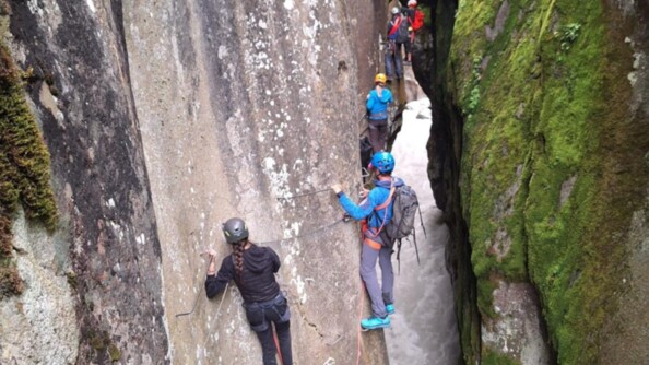 Cinq participants au séjour escalade une paroi rocheuse lors de la via-ferrata. Sous leur pied, se trouve un cours d'eau creusant un sillage étroit dans la roche.