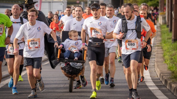 Des coureurs, vêtus d'un T-shirt blanc avec un numéro sur le ventre, courent sur une avenue pavée, en poussant le fauteuil roulant d'un jeune garçon. On aperçoit un pont en arrière plan.