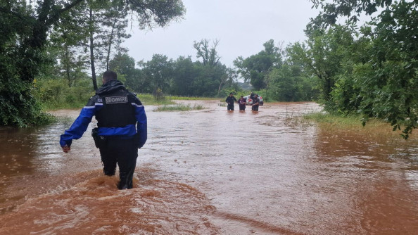 Un gendarme, vu de dos, progresse à pied dans les eaux boueuses, en direction de trois personnes également dans l'eau jusqu'aux genoux.