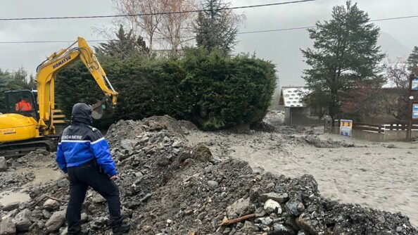 Un gendarme de dos, vêtu d'un blouson de pluie, capuche sur la tête, sur une digue de fortune érigée à proximité d'un fleuve de boue dévalant au milieu des habitations. En arrière-plan, à gauche, une tractopelle en action prolonge la digue.