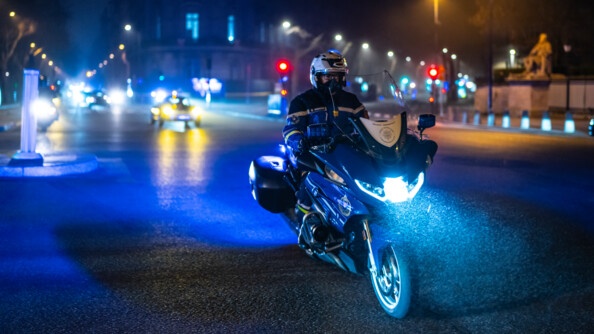 Un gendarme en moto de nuit devant des véhicules de collection