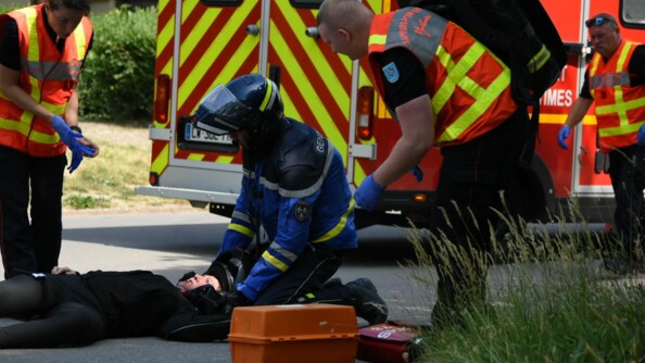 Reconstitution d'un accident de moto : un motocycliste de la gendarmerie est agenouillé auprès d'un motard à terre. Ils sont entourés par deux sapeurs-pompiers ; un troisième est en arrière-plan, à côté du camion du SDIS.