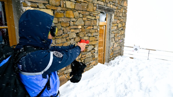 un gendarme, de dos, à sa gauche le mur d'un châlet, pointe une cible avec un pistolet rouge d'exercice