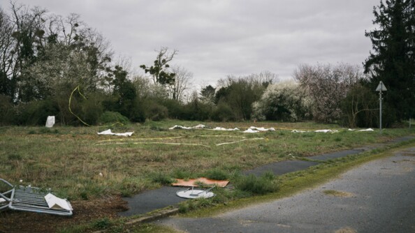 Paysage du lieu de l'exercice. Au sol des zones délimitées par des rubans de gendarmerie. Une dizaine de draps mortuaires au sol pour illustrer le corps des victimes. Tout cela est situé dans de l'herbe, un peu boueuse et humide.