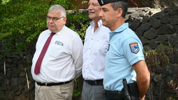 Deux hommes en civil et un colonel de gendarmerie en tenue d'intervention, côte à côte et regardant vers la gauche de l'image, devant un muret de pierre parsemé de végétation