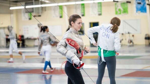 La sabreuse Manon Brunet à l'entraînement à Orléans, masque sous le bras droit et sabre dans la main droite. Au second plan, d'autres sabreuses dont une chinoise de dos.