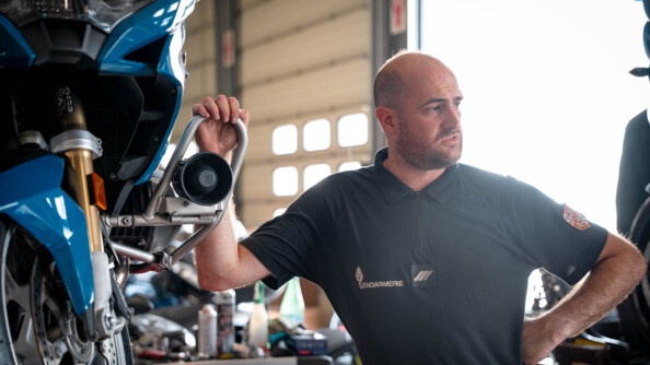 Dans un atelier de réparation automobile, un mécanicien se tient à côté d'une moto bleue de la gendarmerie, sa main droite posée sur le véhicule. L'homme est vêtu d'un t-shirt noir, portant sur la poirtrine l'inscription Gendarmerie.