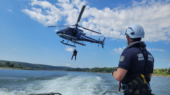 Un h&eacute;licopt&egrave;re bleu marine de la Gendarmerie nationale effectue une op&eacute;ration d&rsquo;h&eacute;litreuillage au-dessus d&rsquo;un plan d&rsquo;eau. Un gendarme est suspendu &agrave; un c&acirc;ble, en cours de descente depuis l&rsquo;h&eacute;licopt&egrave;re. Au premier plan, un autre gendarme, &eacute;quip&eacute; d&rsquo;un casque et d&rsquo;un harnais, observe la sc&egrave;ne depuis un bateau en mouvement. En arri&egrave;re-plan, on distingue un paysage de collines verdoyantes sous un ciel bleu parsem&eacute; de nuages blancs.