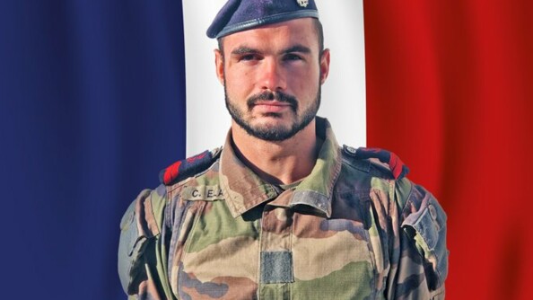 Un jeune militaire brun, yeux marrons, portant une barbe, pose en tenue kakie devant un drapeau bleu blanc rouge recouvrant intégralement le fond de l'image.
