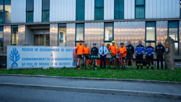 Une jeune femme cycliste, pose entourée de deux cysclistes avec un t-shit ornage sur sa droite, deux autres cyclistes également avec un t-shit ornage sur sa gauche, ainsi que cinq autres personnes (gendarmes). Derrière eux se trouve un bâtiment, et sur leur droite un grand panneau portant l'inscription Région de gendarmerie de Bretagne