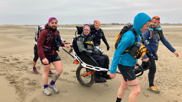 Un jeune homme sur une joëlette, tracté par deux personnes à l'avant, et poussé par deux personnes à l'arrière, avance, souriant, sur une plage. Le ciel est gris.