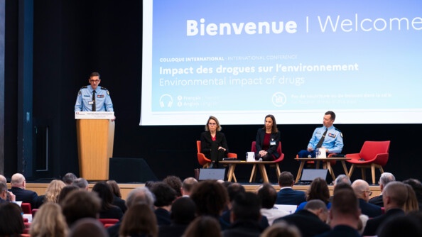 Photo d'un intervenant du colloque, qui s'exprime au micro devant l'assembl&eacute;e, debout derri&egrave;re un pupitre en bois. Derri&egrave;re lui on aper&ccedil;oit un grand &eacute;cran sur lequel est inscrit le titre du colloque "Impact des drogues sur l'environnement", ainsi que la mention "Bienvenue"