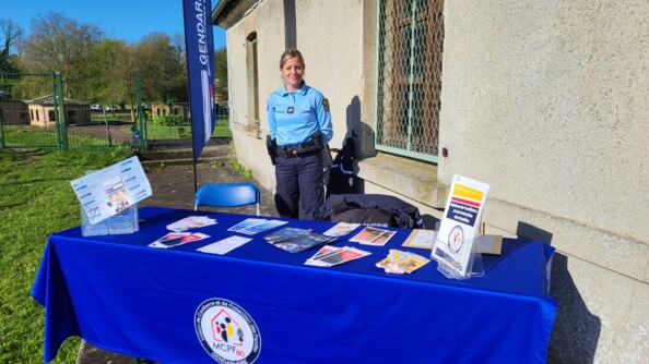 Une femme gendarme se tient bras le dos derrière une table recouverte d'une nappe bleu roi surmontée du logo de la Maison de protection des familles. La table est recouverte de prospectus. En arrière plan à droite un batiment gris clair et à gauche de la verdure et des petits bâtiments anciens