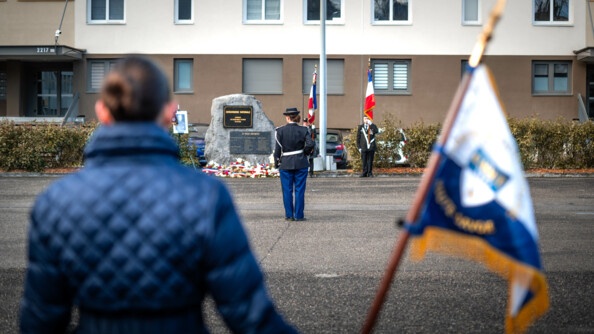 En flou de dos, une femme au manteau bleu tient un fanion blanc jaune et bleu, au fond des gendarmes en tenue de cérémonie et drapeau devant une stèle