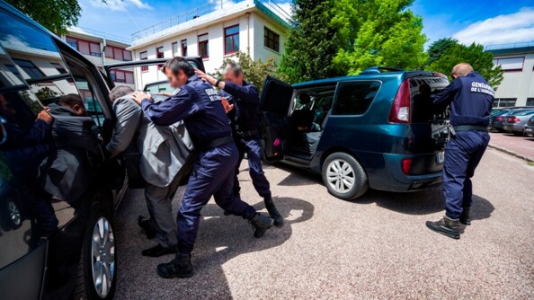 Deux gendames enjoignent un homme en tenue civile d'entrer dans une voiture. L'un deux a ses deux mains posées sur le dos du civil, qui se tient courbé
