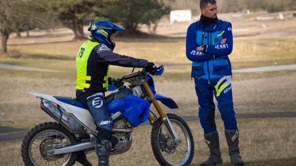 un motard de la gendarmerie sur une moto cross à l'arrêt, de profil, avec devant lui, à droite, un instructeur debout. Le tout sur un terrain herbeux sauvage avec quelques arbres