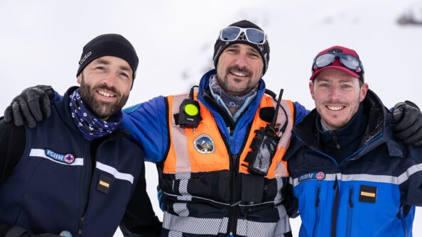 Dans un paysage enneigé, trois hommes souriants, vêtus en tenue de montagne, se tiennent debout, en se tenant pas les épaules et la taille. Tous les trois regardent l'objectif.