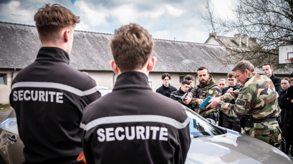 Cours d'intervention professionnelle au profit des lycéens - au premier plan vers la gauche, deux jeunes hommes de dos vêtus d'un pull noir floqué "sécurité" en blanc. Au second plan, derrière une voiture Renault grise, deux militaires effectuent une démonstration d'un contrôle avec arme factice. Ils sont entourés d'une dizaine de jeunes qui suivent la démonstration. En arrière-plan, un bâtiment en longueur recouvert d'ardoises.