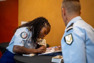 Une femme en tenue de gendarme signe une carte sur une tabe lors d'une séance de dédicace
