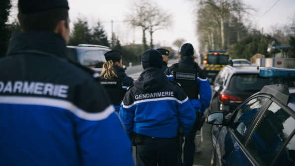 5 gendarmes de dos dans une rue. Sur les côtés quelques voitures garées.
