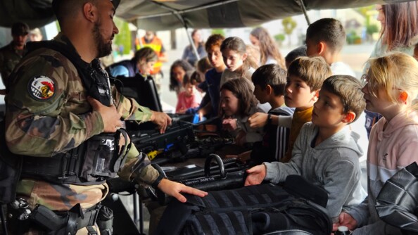 Un militaire en tenue derrière un stand sur lequel se trouve du matériel (gilet pare-balle, armess...). De jeunes enfants âgés de 6 à 8 ans se trouve face au gendarme. Un jeune garçon saisit dans sa main droite le gilmet pare-balle.