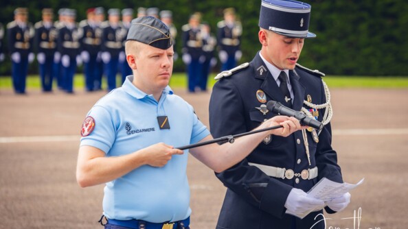 un élève-gendarme lit un texte tandis que d'un autre élève, en tenue d'intervention, lui tend le micro