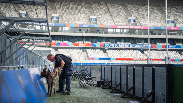 Un gendarme en noir sécurise un stade avec son chien , un malinois. La mention "coupe du monde de rugby" et "bordeau métropole" recouvre les tribunes