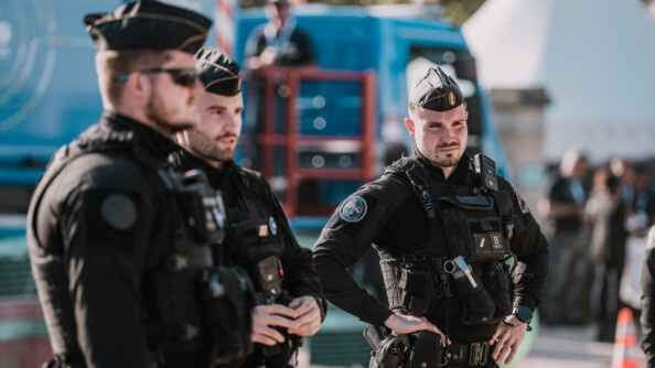 Trois gendarmes mobiles de face et de profil et en fond, flou, un camion régie de la cérémonie d'Omaha Beach.