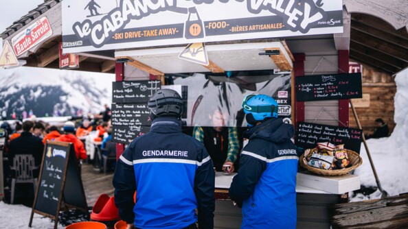 Deux gendarmes se tiennent debout, côte à côte, de dos, au niveau d'un petit cananon en bois vendant de la nourriture et des boissons. On peut lire sur l'inscription "Le cabanon d'happy valley". On aperçoit en arriière plan de nombreux skieurs installés en terrasse, ainsi que la montagne enneigée.