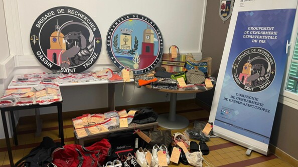 De multiples objets sous scellés, placés dans des sachets, posés sur des tables. Au sol, des paires de chaussures et des sacs. Contre le mur, à droite, le kakémono de la compagnie et contre le mur, au fond, l'écusson de la B.R. et celui de la gendarmerie de Saint-Tropez.