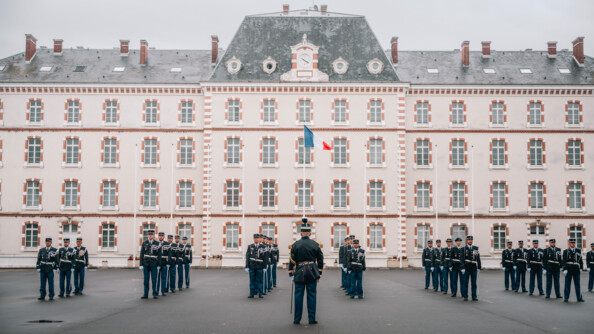 Des gendarmes en tenue de cérémonie en colonne par 6 sur la place d'armes devant un bâtiment. Au milieu,  de dos, un officier en tenue de tradition