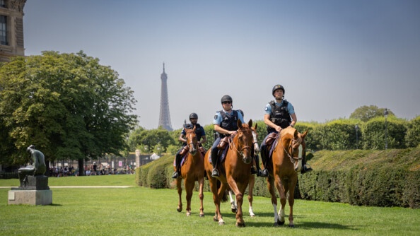 Quatre cavaliers, marchant deux par deux, sur l'herbe, devant des haies et arbres, ainsi qu'une statue à gauche. En fond, la Tour Eiffel